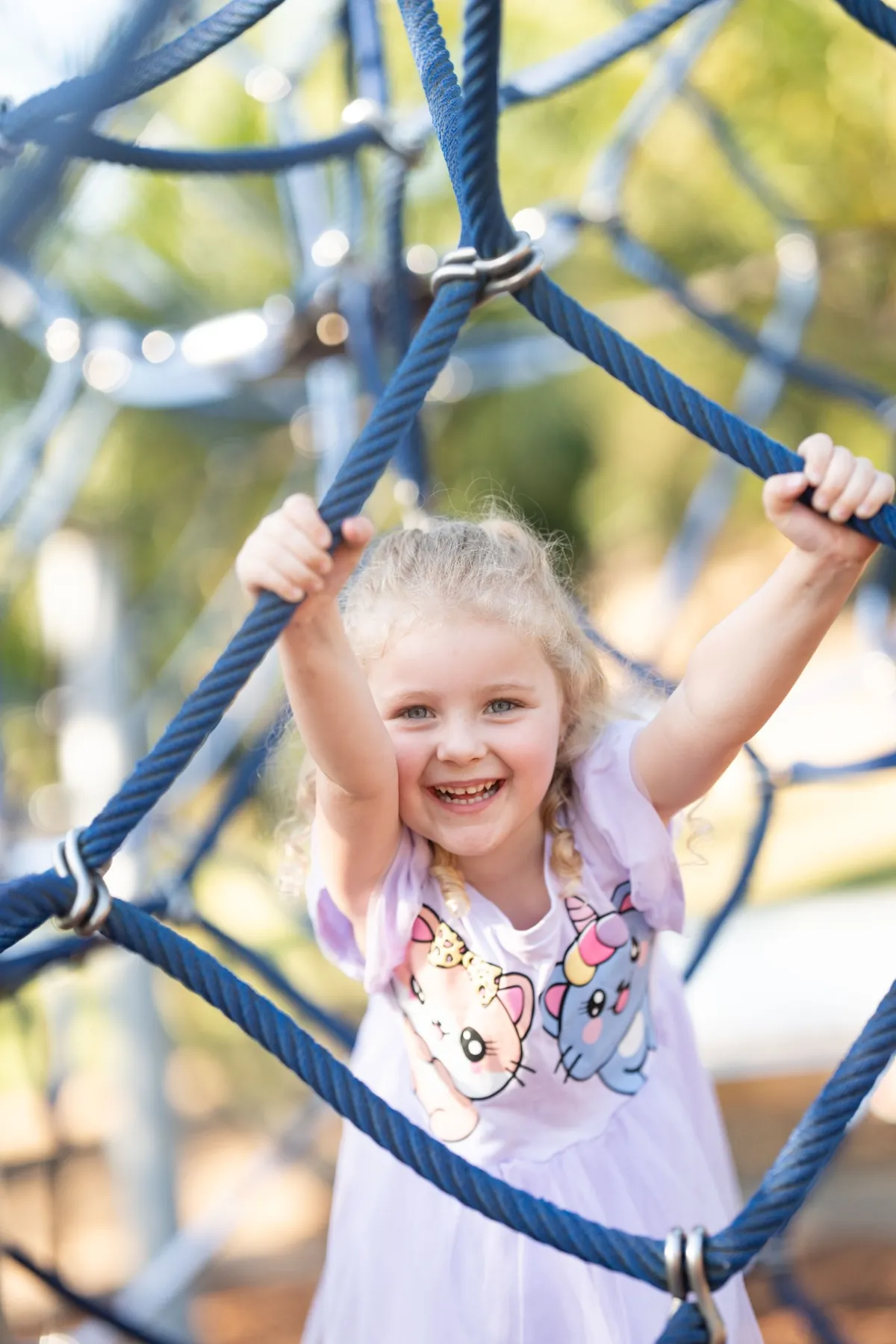 Young girl smiling while climbing blue rope playground equipment in Brisbane QLD, photographed by Little Star Photography.