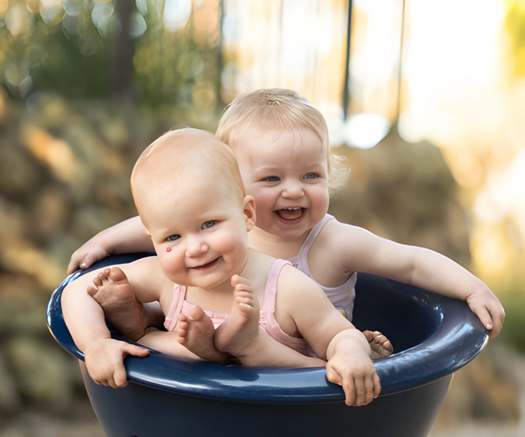 Twin babies laughing together while sitting in a blue bowl outdoors in Ipswich, photographed by Little Star Photography.