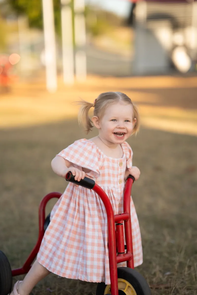 Laughing toddler girl riding a red trike in a Brisbane QLD park, captured by Little Star Photography.