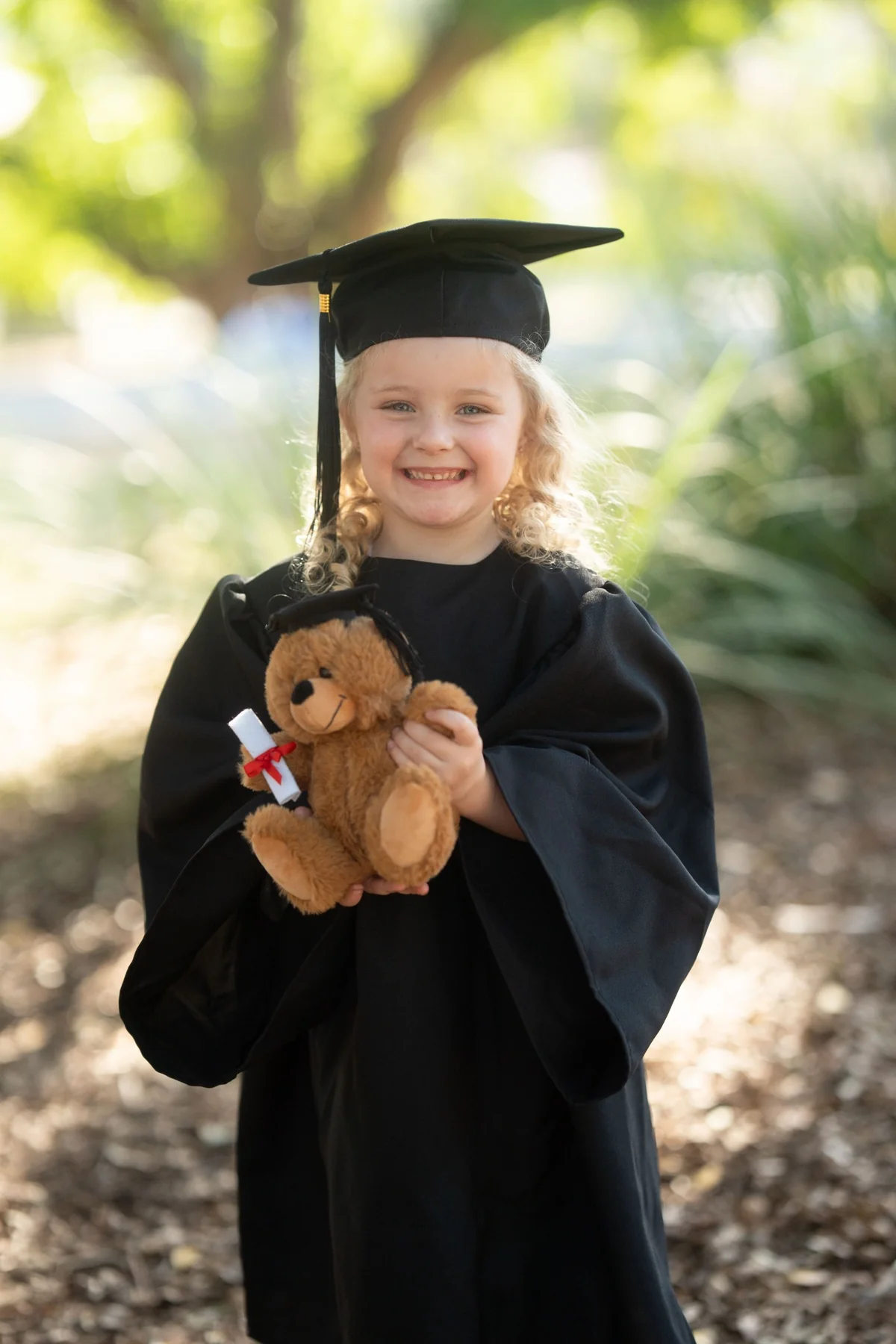Smiling preschool graduate holding a teddy bear dressed in a graduation cap photographed by Little Star Photography in Brisbane QLD.