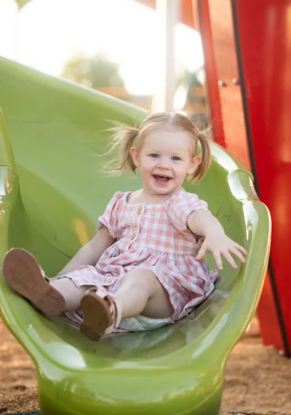 Girl with pigtails laughing while sliding down a green slide at a playground photographed by Little Star Photography in Brisbane QLD.