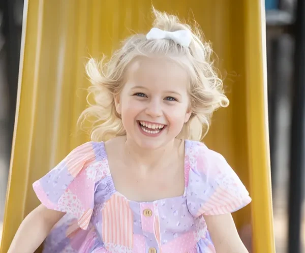 Smiling girl in a pastel dress sliding down a yellow playground slide.