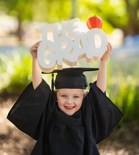 Child in graduation gown holding a “The Grad” sign above their head.