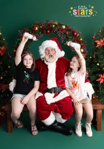 Santa sitting with two girls raising milk bottles during a fun Ipswich Christmas photoshoot by Little Star Photography.