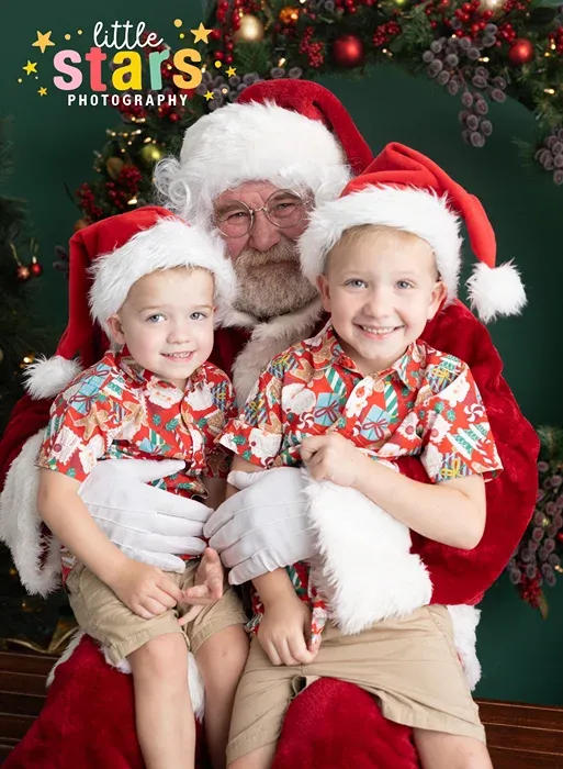 Santa Claus posing with two young boys wearing Christmas shirts and Santa hats.