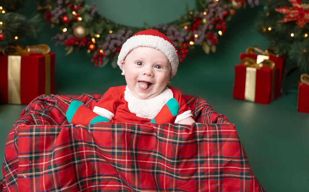 Happy baby leaning over a red plaid blanket in front of Christmas decorations during a Christmas baby portrait.