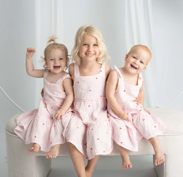 Three young siblings in matching pink dresses sitting under a floral arch during a siblings studio portrait.
