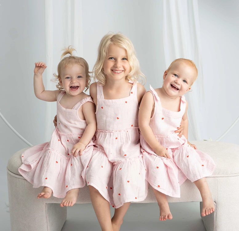 Three young siblings in matching pink dresses sitting under a floral arch during a siblings studio portrait.