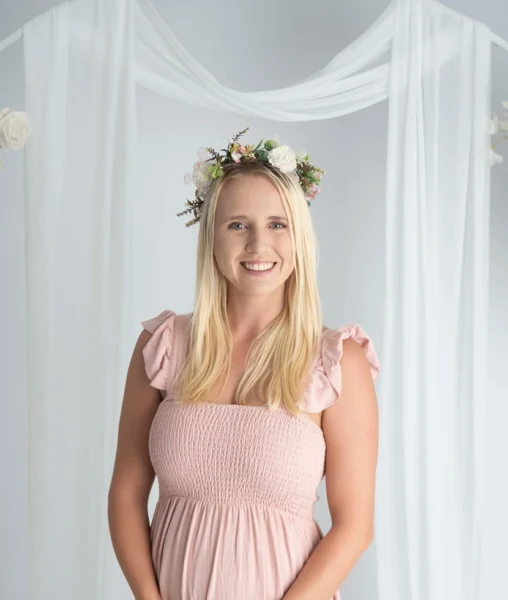 Smiling woman wearing a pink dress and floral crown against a soft white studio backdrop.