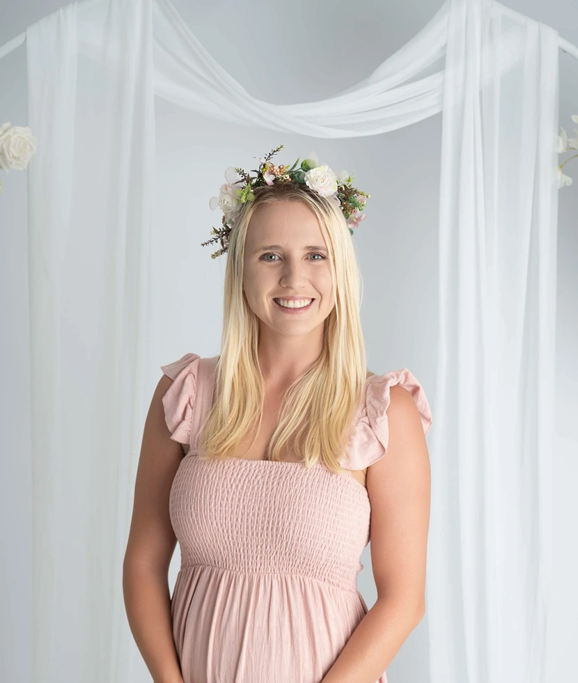 Smiling woman wearing a pink dress and floral crown against a soft white studio backdrop.