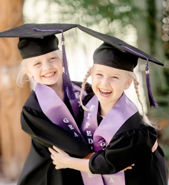 Two young girls in graduation gowns and caps hugging and smiling at their kindergarten graduation ceremony in Queensland.