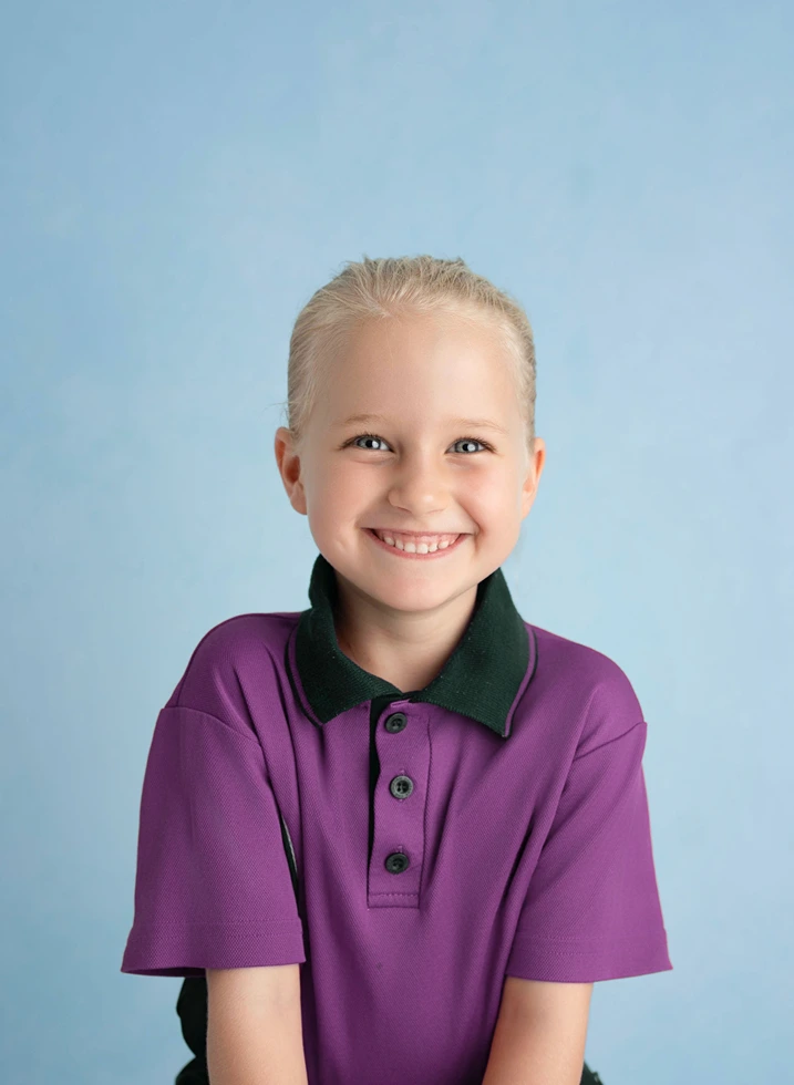 Smiling primary school girl in purple school uniform against a soft blue studio background.