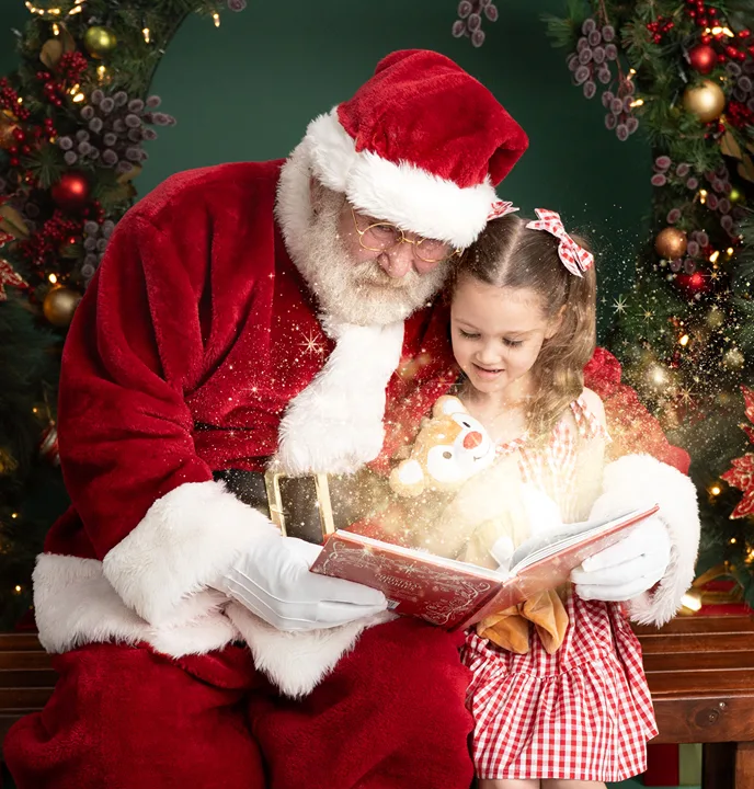 Santa reading a glowing Christmas book with a young girl during a Santa magic portrait session.