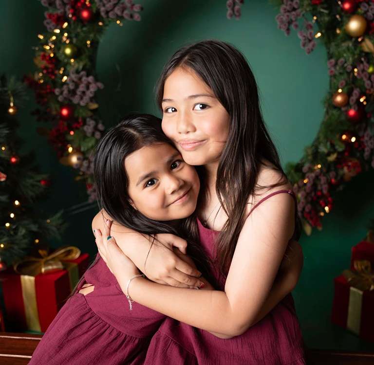 Two young sisters in red dresses hugging in front of Christmas decorations during a Christmas siblings portrait.