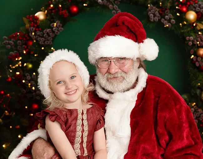 Young girl sitting with Santa Claus in front of a festive Christmas wreath backdrop.