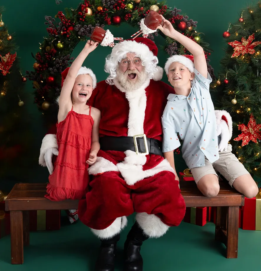 Smiling siblings holding a Santa toy while sitting on a bench beside a Christmas tree in a studio, photographed in Brisbane by Little Stars Photography.